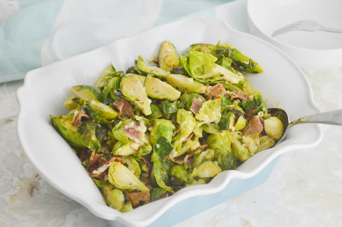 A white dish filled with Keto Bacon Brussels Sprouts Casserole mixed with crispy bacon pieces. A spoon rests on the side of the dish, and a fork is visible in the background. The dish is placed on a light-colored surface.
