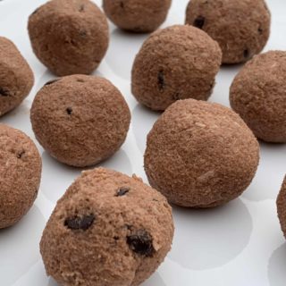 Close-up of several round, Keto Chocolate Coconut Cookie Dough Bites arranged on a white plate, ready for baking or as a snack. The texture looks soft and slightly granular, with visible small chocolate chips scattered throughout.
