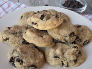 A stack of homemade Keto chocolate chip cookies with some scattered loose chocolate chips on a white plate, suggesting a cozy, freshly-baked low-carb treat.