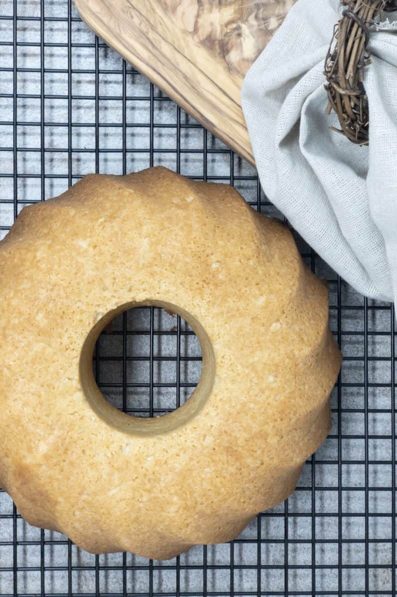 A freshly baked Keto Almond Maple Cake on a black wire cooling rack, surrounded by a wooden cutting board and a folded beige cloth with a wreath detail. The cake has a smooth, golden-brown surface.