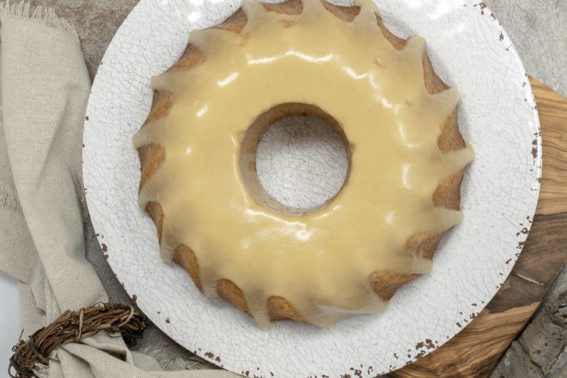 A glazed Keto Almond Maple Cake on a white plate and a wooden board. The top half shows the whole cake, while the bottom half features a slice on a white plate. Text reads "Keto Almond Maple Cake." A whisk lies beside the board.