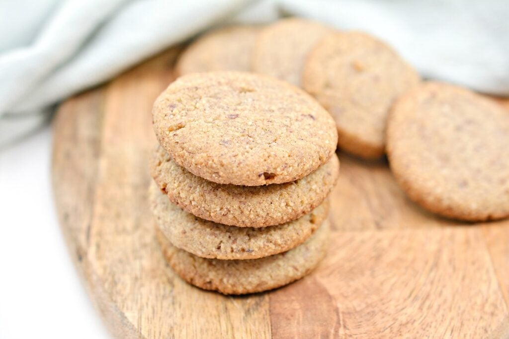 A stack of five round, golden-brown Keto Ginger Snaps on a wooden cutting board, with a few more cookies lying flat in the background. A soft, light blue cloth is partially visible in the upper left corner.
