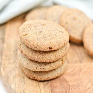 A stack of five round, golden-brown Keto Ginger Snaps on a wooden cutting board, with a few more cookies lying flat in the background. A soft, light blue cloth is partially visible in the upper left corner.