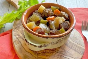 A ceramic bowl filled with a hearty Beef and Veggie Stew, featuring chunks of beef, carrots, potatoes, and celery. The bowl sits on a wooden board with a red napkin beneath it. A fork and a celery stalk are nearby.