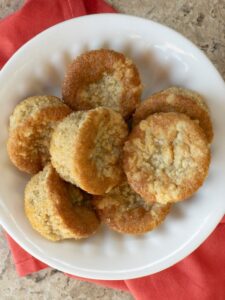 A plate of golden-brown Keto Cinnamon Crumb Cake Muffins are arranged on a white dish, placed on a red cloth. The muffins have a slightly crumbly texture and appear freshly baked.