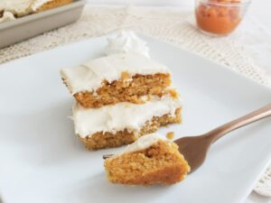 Two stacked Keto Pumpkin Bars, each coated in white icing, are served on a white plate. A fork is piercing a piece of the cake on the plate. In the background, there is a dollop of whipped cream and a corner of another dish visible.