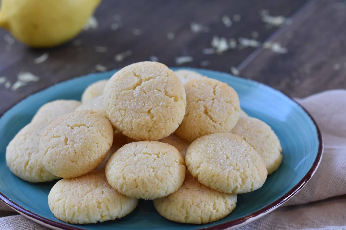 A plate of freshly baked, soft Keto Lemon Cookies piled up on a blue plate, displaying a slightly cracked texture and soft yellow color, with a blurred lemon in the background.