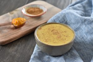 A bowl of creamy, Keto Pumpkin Pudding with a sprinkle of pumpkin pie spice sits on a wooden serving board. A small spoon lies beside it, and a blue cloth is draped around the board. In the background, there's a small dish with a brown spice. The setting is on a dark wooden table.