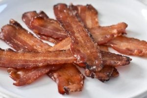 A white plate with several crispy, cooked Keto Maple Bacon strips placed on a light fabric surface.