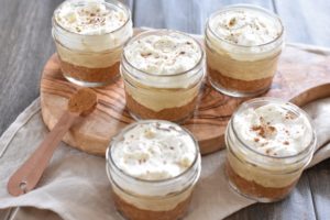 Five small glass jars filled with layered Keto Pumpkin Mousse sit on a wooden surface. Each jar contains a crumbly base, a creamy middle layer, and a topping of whipped cream sprinkled with a hint of cinnamon.