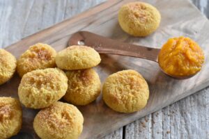 A wooden board with several Keto Pumpkin Spice Cookies, lightly dusted with sugar. A copper measuring spoon containing pumpkin purée rests beside the cookies on a rustic wooden surface.
