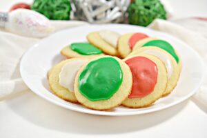 A white plate with nine Keto Christmas Cookies frosted in red, green, and white icing. The plate is surrounded by decorative green and silver spheres on a light-colored cloth.