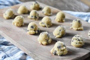 Scoops of Keto Cookie Dough sit on a wooden cutting board. An ice cream scoop with cookie dough is visible in the background. The setting appears to be a kitchen counter.