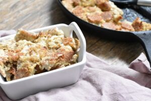 A white ceramic dish filled with baked keto bread pudding sits on a wooden table. In the background, a black skillet holds more stuffing. A beige cloth is partially visible beside the dish.