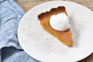 A slice of Keto Pumpkin Pie with a dollop of whipped cream on top, served on a white plate. The pie has a golden-brown crust and is lightly sprinkled with cinnamon. The plate is placed on a rustic wooden surface with a blue cloth napkin beside it.