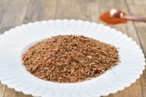 A white plate holds Keto Fajita Seasoning on a wooden surface. In the background, a small copper spoon with additional spices is visible.