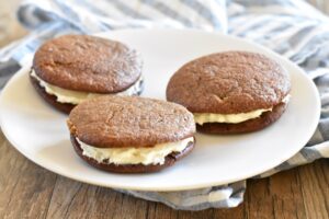 Three Gluten Free Gingerbread Whoopie Pies with cream filling sit on a white plate, placed on a wooden table with a striped cloth in the background.