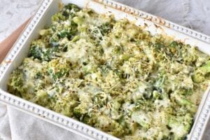 A casserole dish filled with baked Keto Broccoli Casserole, topped with melted cheese and sprinkled with herbs and pepper. The dish has a decorative white border and is placed on a light-colored cloth.