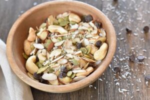 A wooden bowl filled with Healthy Paleo Snack Mix ingredients, including almonds, cashews, pumpkin seeds, coconut flakes, and chocolate chips, on a wooden surface. Scattered around are additional coconut flakes and chocolate chips.