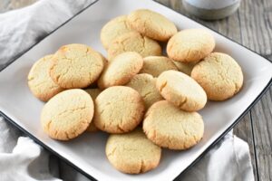 A plate of round, golden-brown Keto Butter Cookies stacked together on a white, square dish. The background surface is wooden, with a dark cloth partially visible under the plate.
