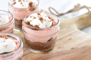 Small glass jars filled with layered Keto Peppermint and Chocolate Pudding Cups, featuring a crumbly base, pink mousse, and whipped cream topping sprinkled with crumbs, placed on a wooden board.