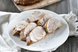 Slices of cooked pork tenderloin arranged on a white plate. The plate is placed on a textured cloth atop a rustic wooden surface, with a cutting board partially visible in the background.