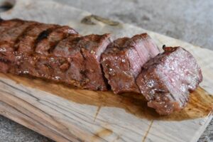 Close-up of a juicy, sliced Grilled Tri Tip on a wooden cutting board. The meat is cooked to medium-rare with a pink interior and a slight outer char, with natural juices pooling around it. A blurred piece of steak is visible in the background.