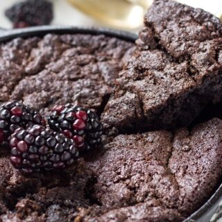 A close-up of a skillet Hemp and Fudge Paleo Brownies topped with fresh blackberries. A slice is being lifted, revealing a rich, fudgy texture. The skillet rests on a striped napkin, and blurred items are visible in the background.