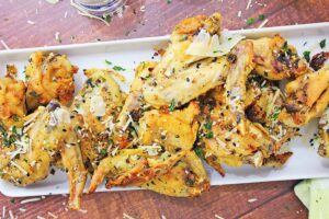 A rectangular plate filled with crispy Keto Garlic Parmesan Wings garnished with sesame seeds, parmesan shavings, and chopped herbs. The wooden table surface and a small glimpse of a green napkin can be seen below the plate.
