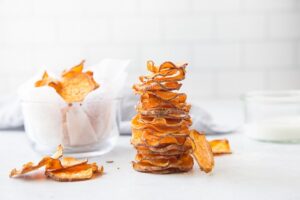 A tall stack of crispy, thinly sliced sweet potato chips stands upright in the center of a light-colored surface. To the left, a glass bowl filled with more chips, lined with parchment paper, is visible. The background is airy and neutral, suggesting a kitchen setting.