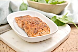 A piece of Air Fryer Keto Blackened Salmon on a white plate, with a side of fresh spinach leaves in the background on a light blue cloth.
