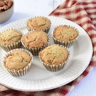 A plate of freshly baked Keto Maple Pecan Muffins on a white wooden surface, accompanied by a bowl of pecans and a red checkered napkin, suggesting a cozy, homemade treat.