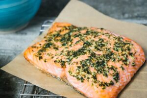 A piece of cooked Keto Lemon Rosemary Salmon fillet topped with herbs and garlic rests on a sheet of parchment paper. The salmon has a bright orange color and a crispy, flavorful topping. It is placed on a wire rack on a kitchen counter.