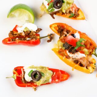 A white plate with colorful Mini Pepper Nachos stuffed with ground meat, topped with sour cream, guacamole, chopped tomatoes, black olives, and fresh herbs. A lime wedge is on the side. Additional stuffed peppers are visible on a tray in the background.