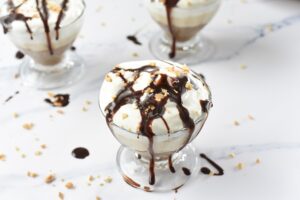 A glass dessert dish filled with layers of creamy Keto Chocolate Peanut Butter Parfait, topped with whipped cream, chocolate sauce drizzle, and chopped nuts. The dish is placed on a white marble surface with scattered bits of nuts and chocolate sauce around it.