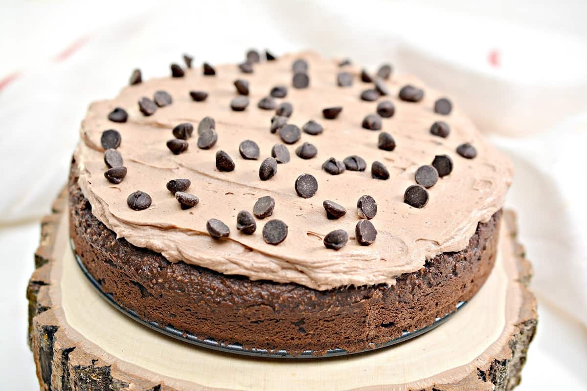 A Keto Chocolate Cake with creamy keto chocolate frosting topped with chocolate chips sits on a rustic wooden stand. The cake has a rich, moist texture and is garnished with numerous chocolate chips spread across the top.