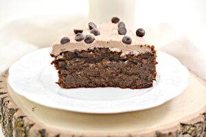 A slice of Keto Chocolate Cake with chocolate frosting and chocolate chips is served on a white plate. A glass of milk sits in the background.