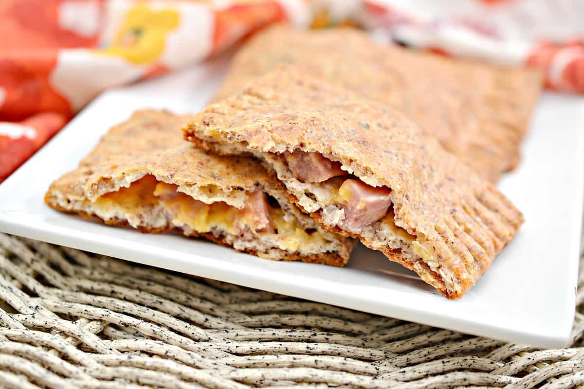 Two golden-brown rectangular Keto Hot Pockets with crimped edges sit on a white square plate. One of the Keto Hot Pockets ha been cut in half. In the background, a colorful floral-patterned cloth is draped. The plate is positioned on a woven, natural-colored surface.