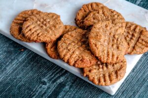 A close-up of several Keto Almond Butter Cookies stacked on a white marble surface. The cookies have a golden-brown color and a crisscross fork pattern on top. The background is a textured dark blue surface.