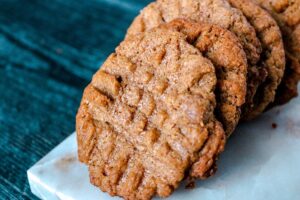 A close-up of several Keto Almond Butter Cookies stacked on a white marble surface. The cookies have a golden-brown color and a crisscross fork pattern on top. The background is a textured dark blue surface.