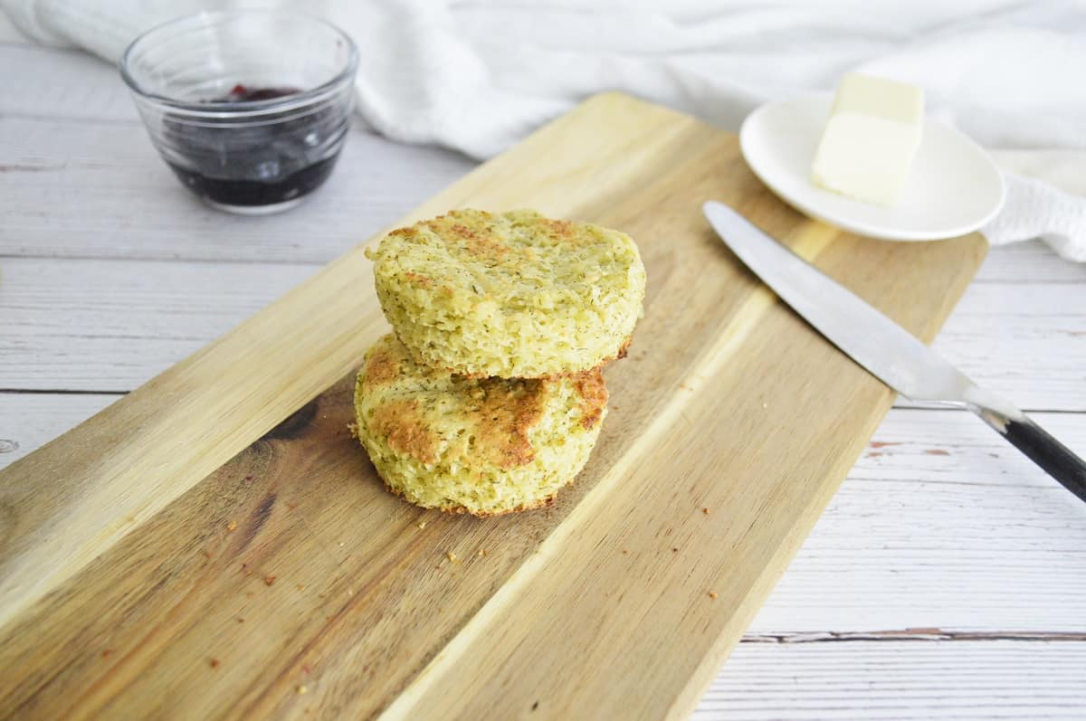 Two freshly baked Keto English Muffins with a golden-brown crust and fluffy interior are stacked on a wooden board. A glass bowl of dark jam and a butter dish are in the background, slightly out of focus. The scene is light and inviting.