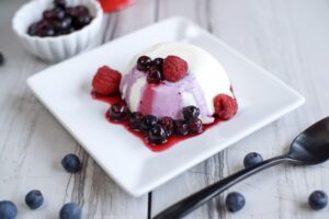 A white and purple Keto Panna Cotta sits on a white rectangular plate, garnished with fresh raspberries and blueberries, and drizzled with berry sauce. A red round container and a small white bowl with more berries are blurred in the background.