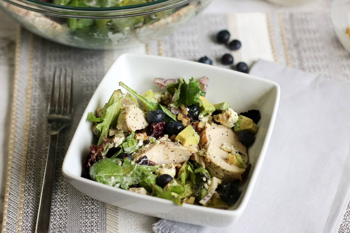 A fresh Keto Avocado Chicken Salad With Blueberries in a white bowl featuring mixed greens, grilled chicken slices, blueberries, avocado chunks, and crumbled cheese. A larger glass bowl with more salad, a dish of crumbled cheese, and a fork are visible in the background, on a table with a striped cloth.