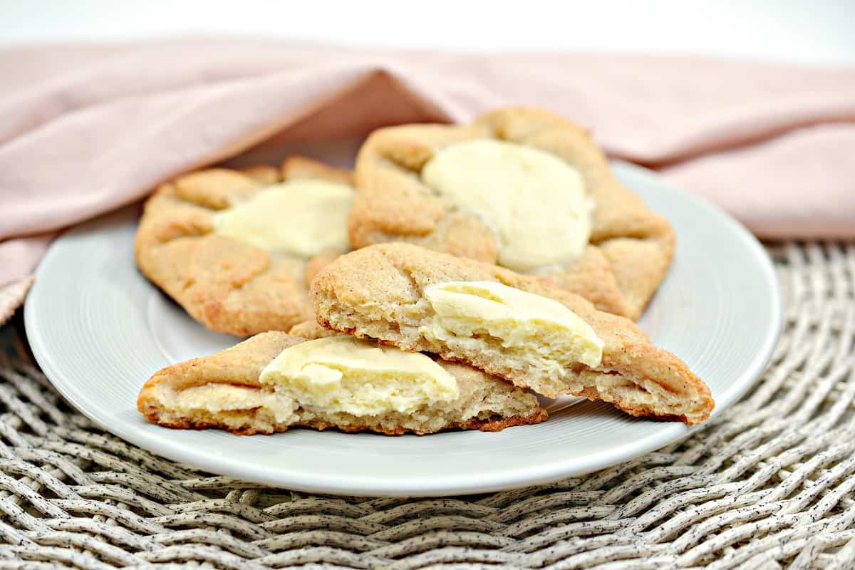 A plate with three cheese Keto Danish pastries. One of the Danishes is broken in half, revealing the creamy cheese filling inside. The pastries are placed on a white plate, with a pink napkin in the background, creating a cozy setting.