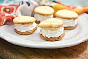 A white plate holds four small Keto Cream Puffs filled with whipped cream, arranged in a circle. The cookies have a golden-brown color and are placed on a wooden surface. In the background, there is a colorful, patterned cloth.
