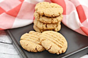 A white plate of five crisscross-patterned Keto Peanut Butter Cookies sits on a dark baking tray. Behind the plate is a red and white checkered cloth. The background is a light-colored wooden surface.