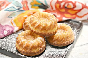 Four small, round, golden-brown Mini Keto Butter Cakes dusted with low-carb powdered sugar are arranged on a black rectangular plate. A vibrant, patterned cloth with floral designs is partially visible in the background.