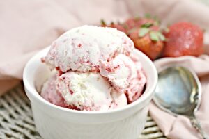 A white bowl filled with scoops of Keto Strawberry Ice cream is placed on a woven mat. In the background, there are fresh strawberries and a metal spoon resting on a pink cloth. The ice cream has visible chunks of strawberries mixed with creamy white vanilla.