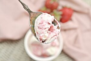 A white bowl filled with scoops of Keto Strawberry Ice cream is placed on a woven mat. In the background, there are fresh strawberries and a metal spoon resting on a pink cloth. The ice cream has visible chunks of strawberries mixed with creamy white vanilla.