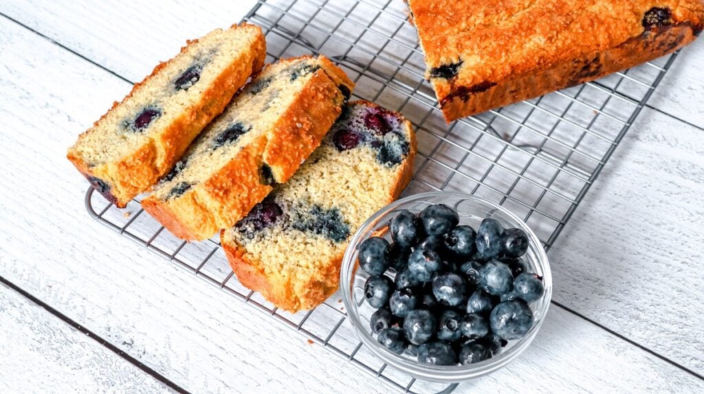A loaf of Keto Blueberry Bread rests on a cooling rack atop a white wooden surface. Three slices are cut from the loaf, revealing blueberries throughout the bread. A small glass bowl filled with fresh blueberries is placed beside the bread.